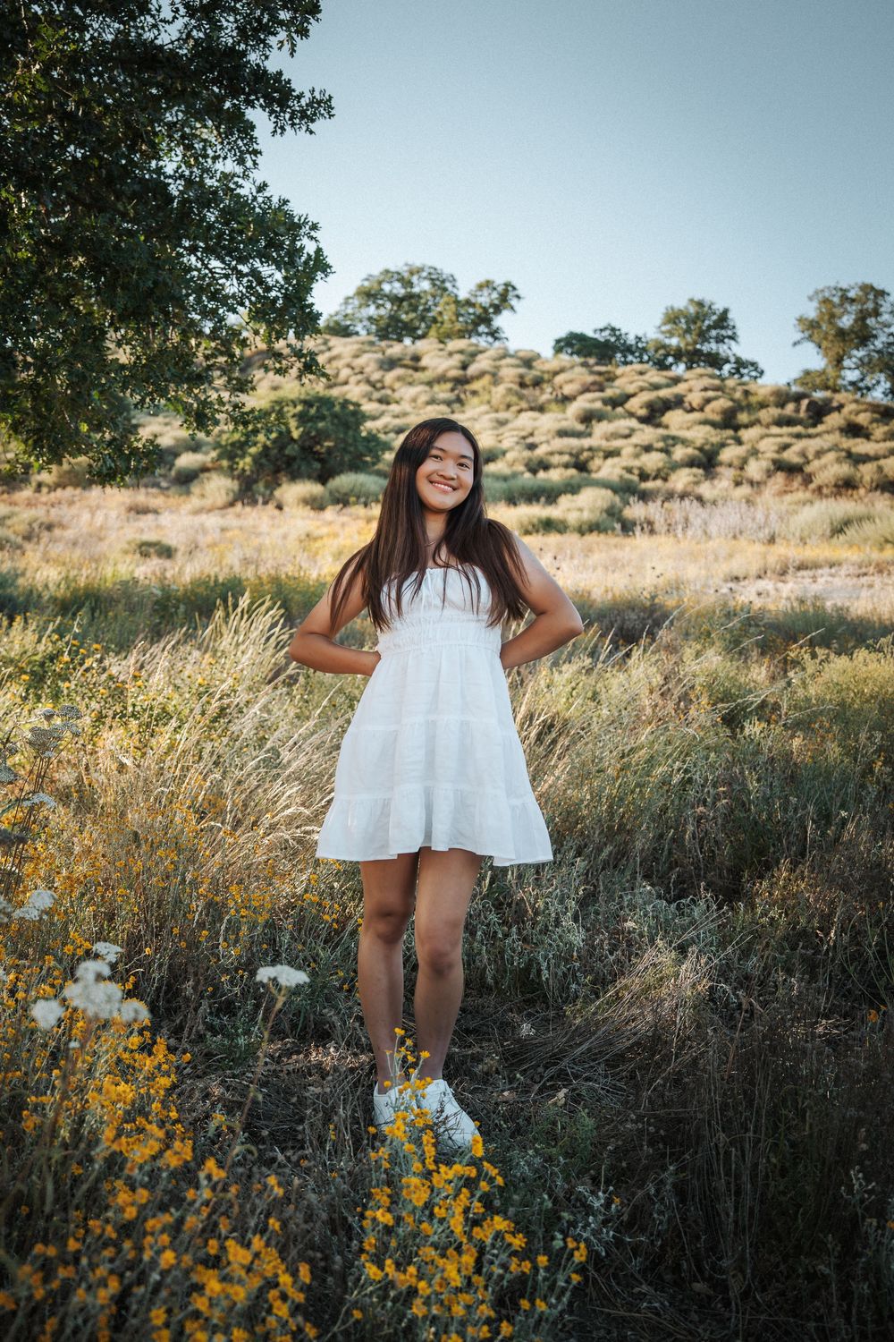 Young woman in white dress smiling in a sunny field with wildflowers and oak trees in the background.