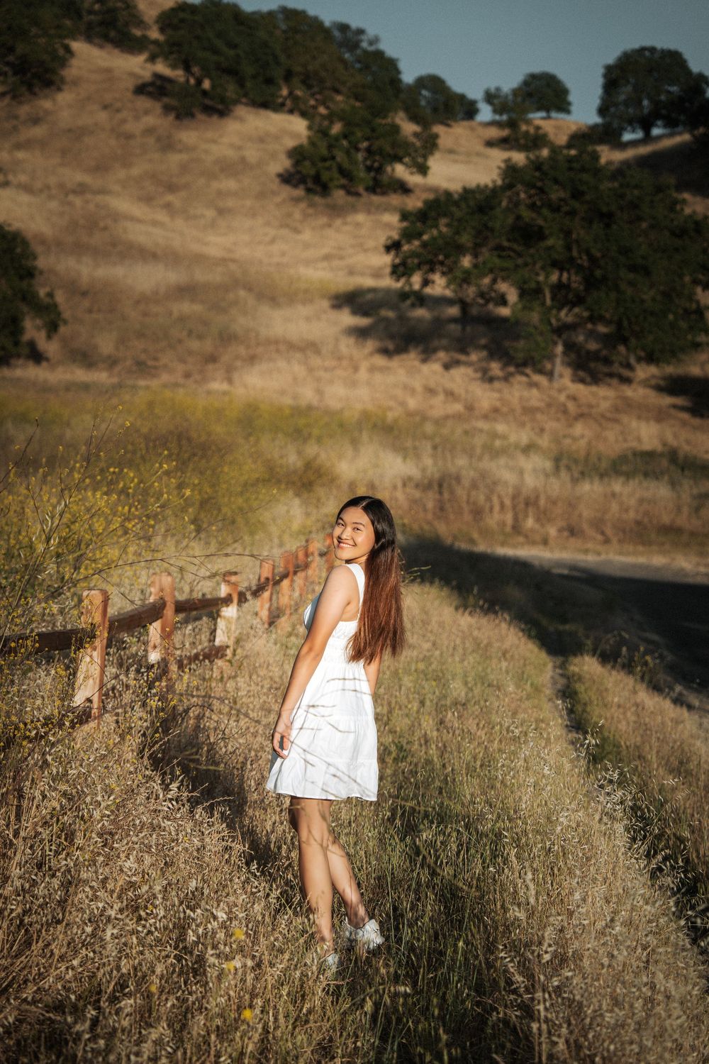 Young woman in white dress walking along a rustic fence in golden California hills during sunset.
