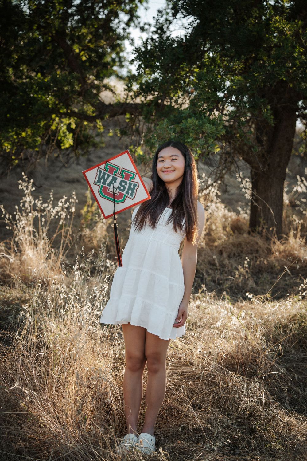 Smiling young woman in white dress holding a WashU pennant in a golden field with trees.