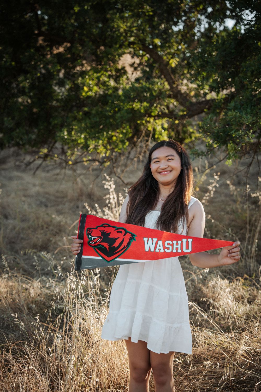 Smiling young woman in white dress holding a red WashU pennant flag outdoors in a golden field.