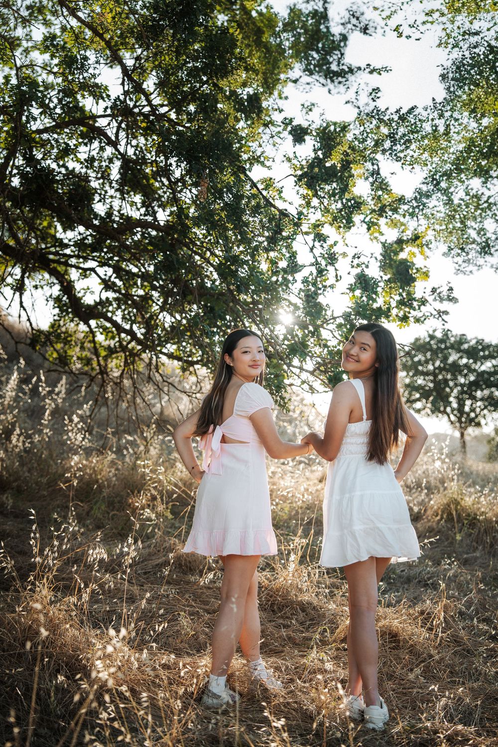 Two young women in white dresses holding hands and posing in a sunlit field with trees.