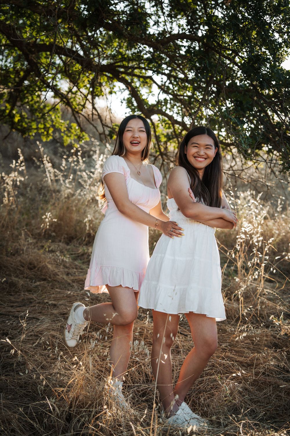 Two young women in white and pink dresses laughing outdoors under a tree in golden hour light.