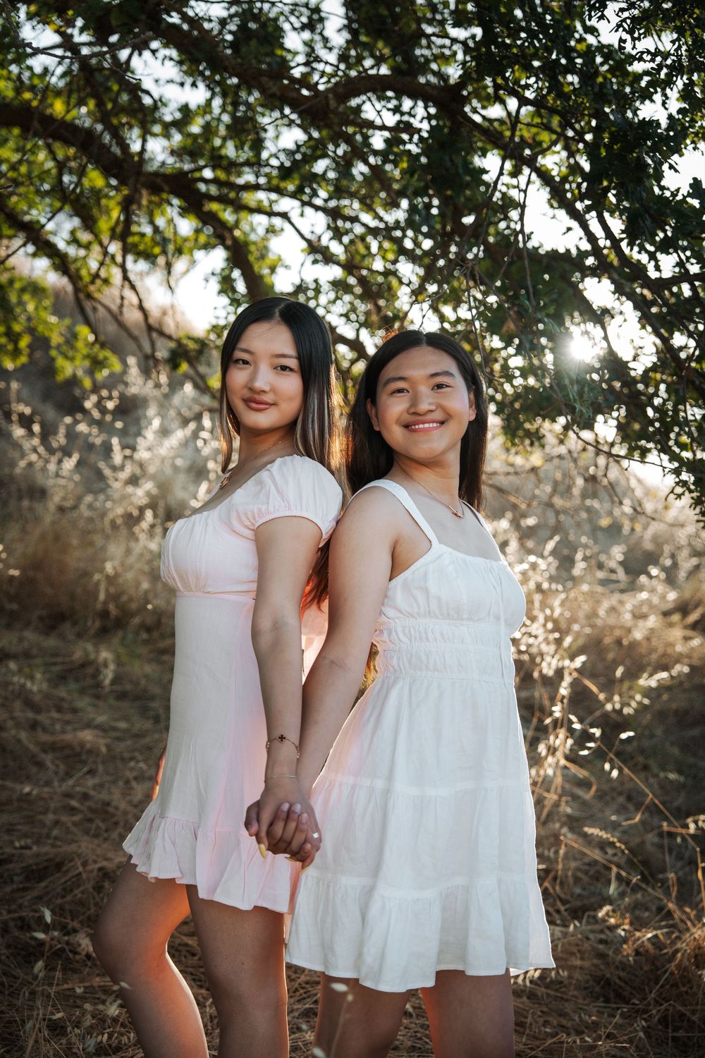 Two young women in white dresses hold hands while posing outdoors under a tree at golden hour.