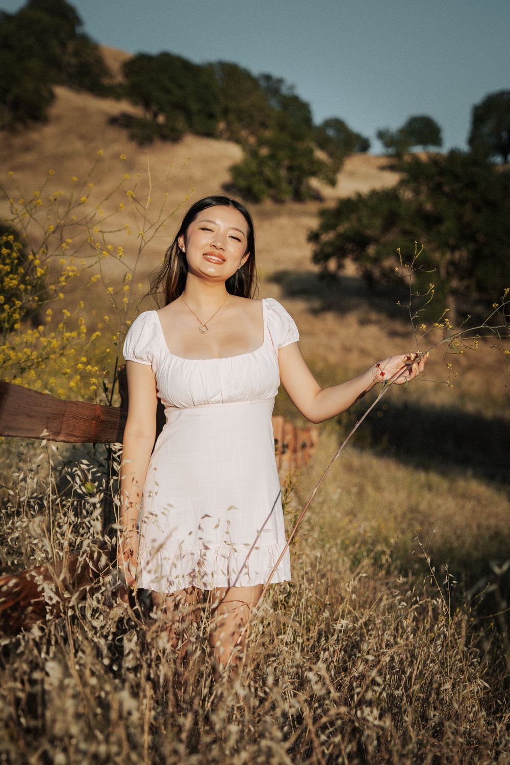 Young woman in white dress standing in golden field with wildflowers and oak trees on a sunny day.