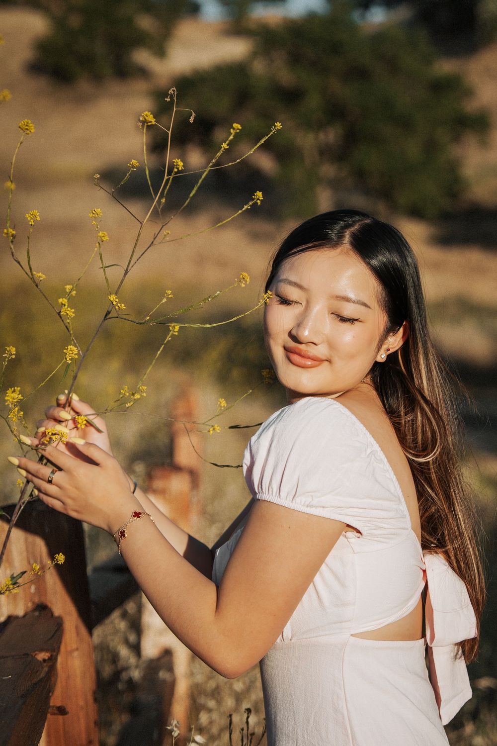Young Asian woman in white dress gently holding yellow wildflower branches outdoors in golden sunlight.