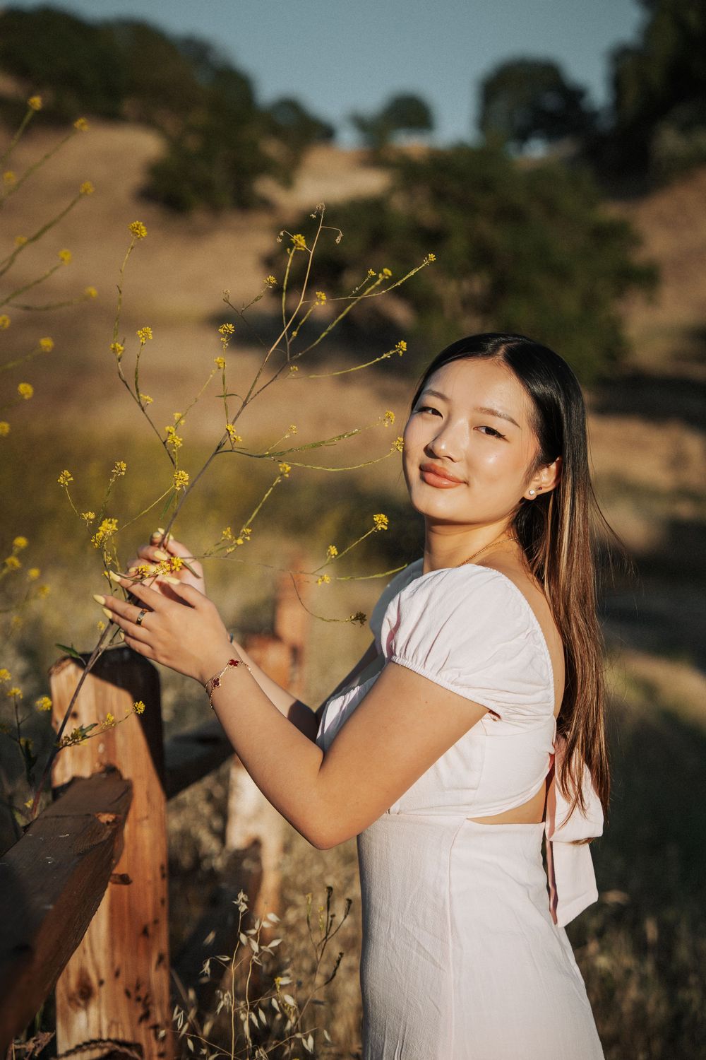 Young woman in white dress smiling while touching yellow wildflowers by a wooden fence in a sunny field.