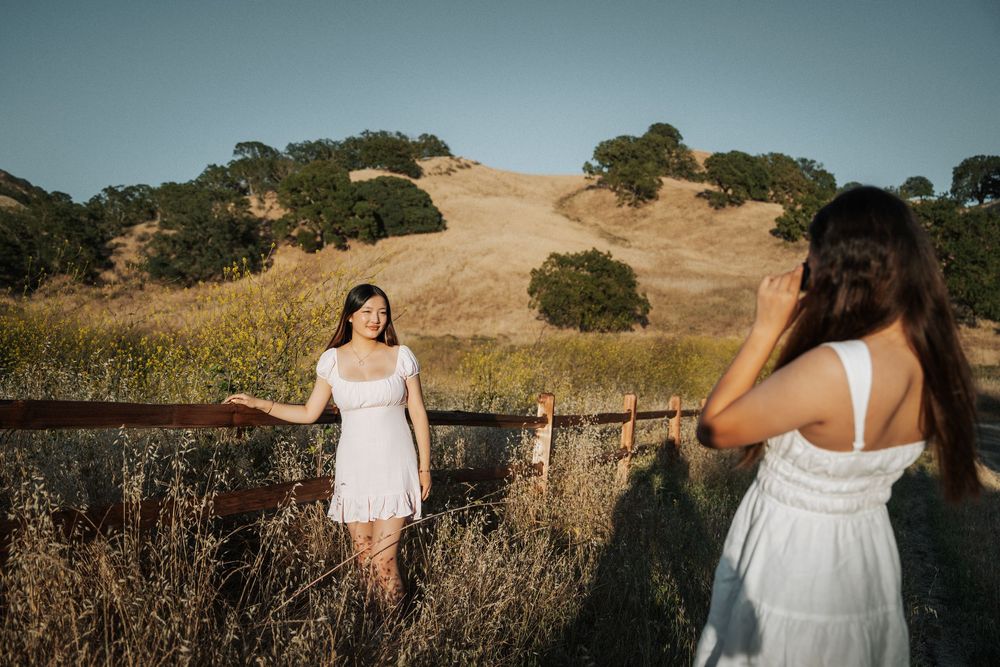 Two young women in white dresses near a wooden fence with golden California hills in the background.