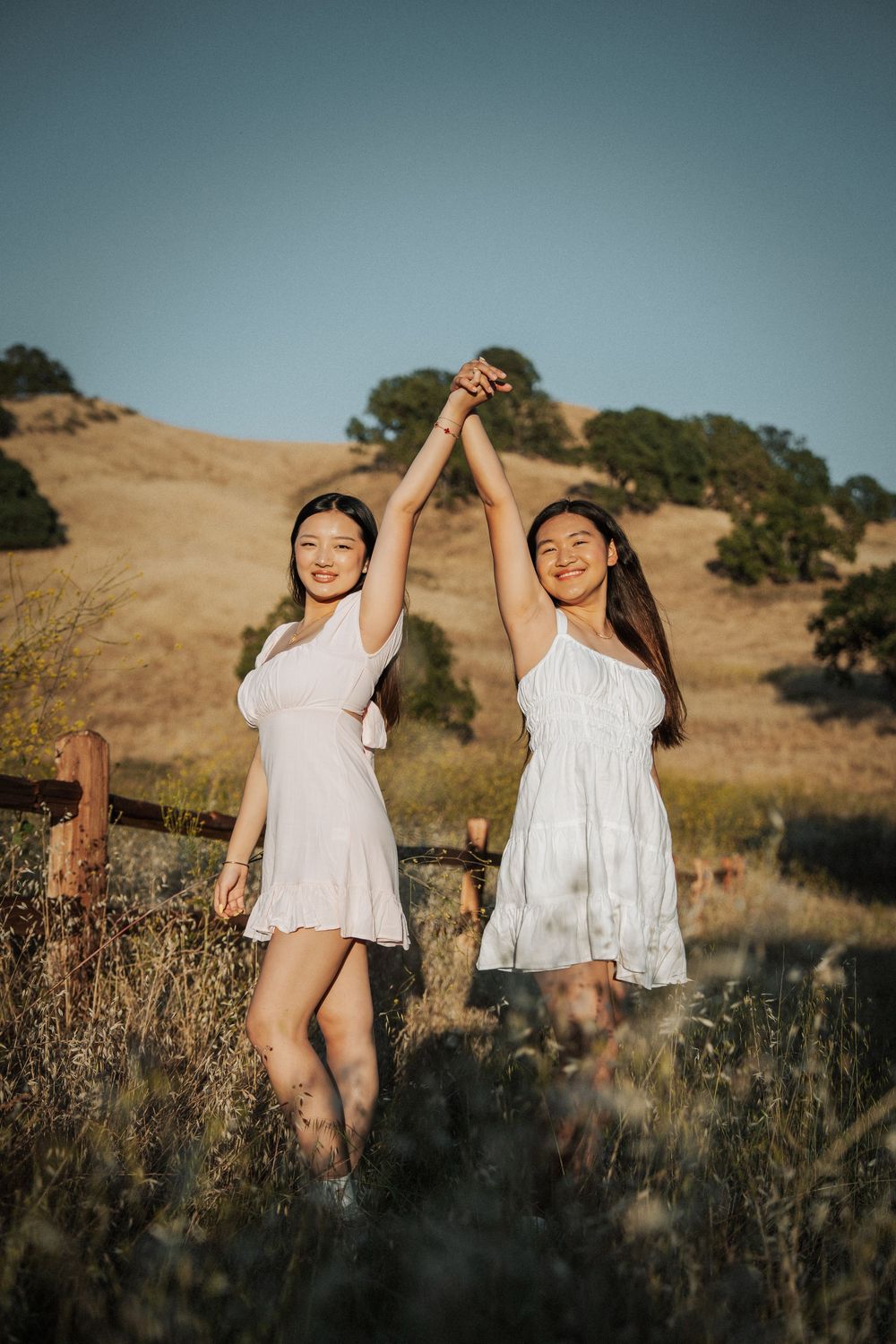 Two smiling young women in white dresses raising hands together in a golden meadow.