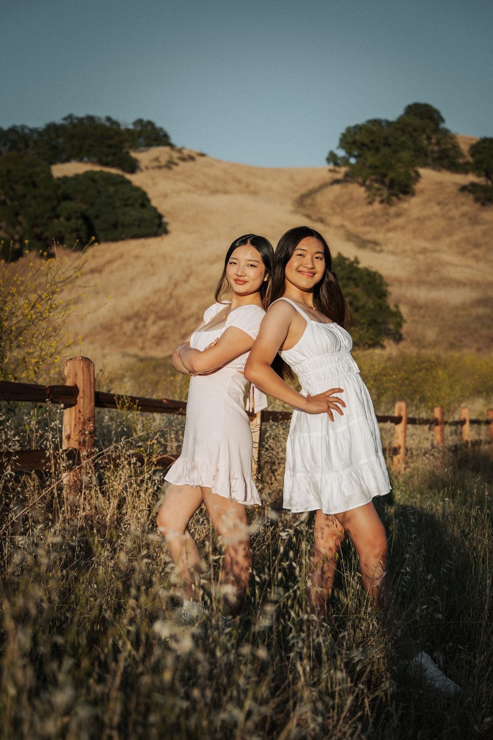 Two young women in white dresses posing back-to-back by a fence in sun-drenched California hills.