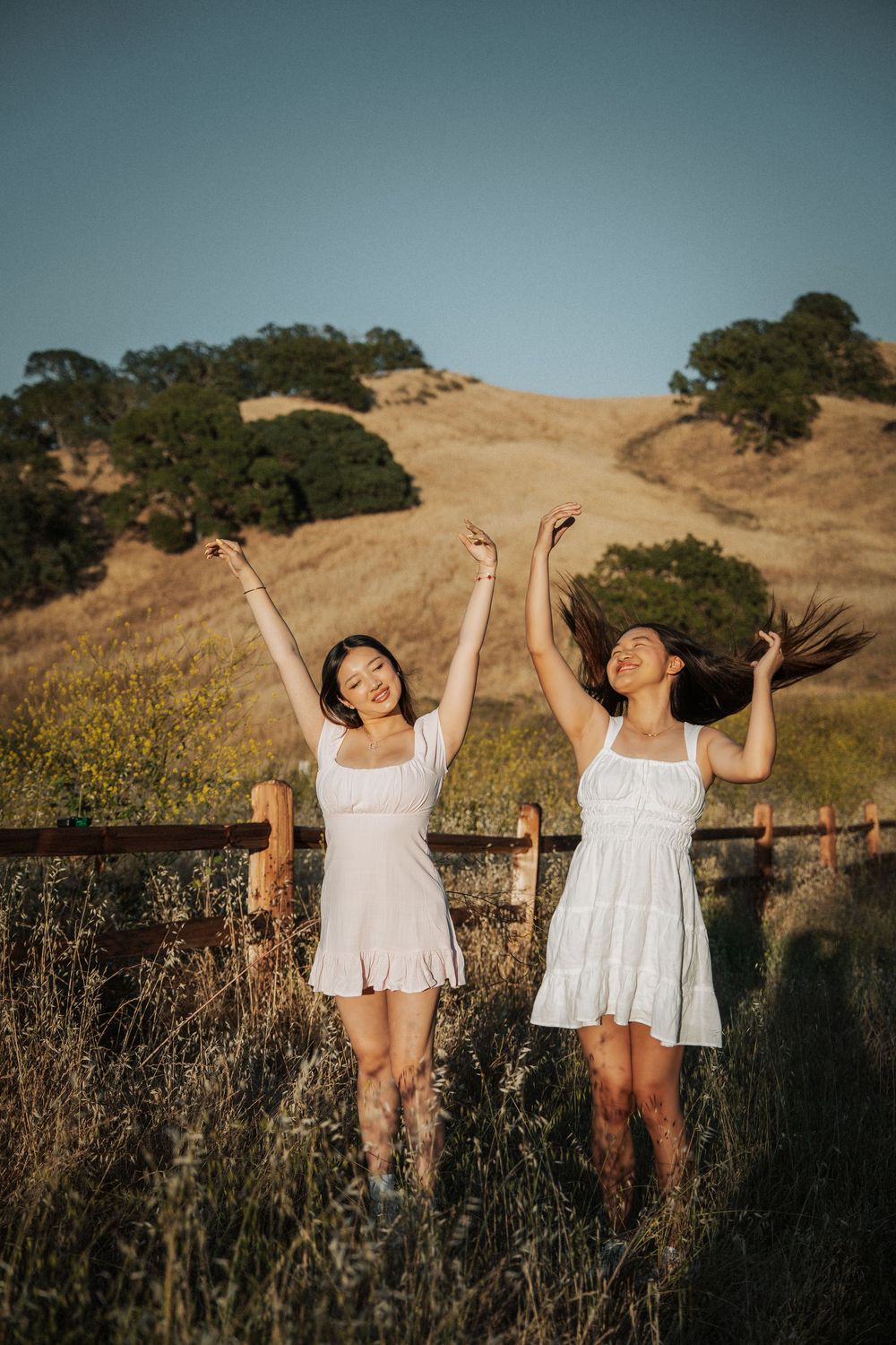 Two smiling young women in white dresses celebrate with arms raised in a sunny golden hillside field.