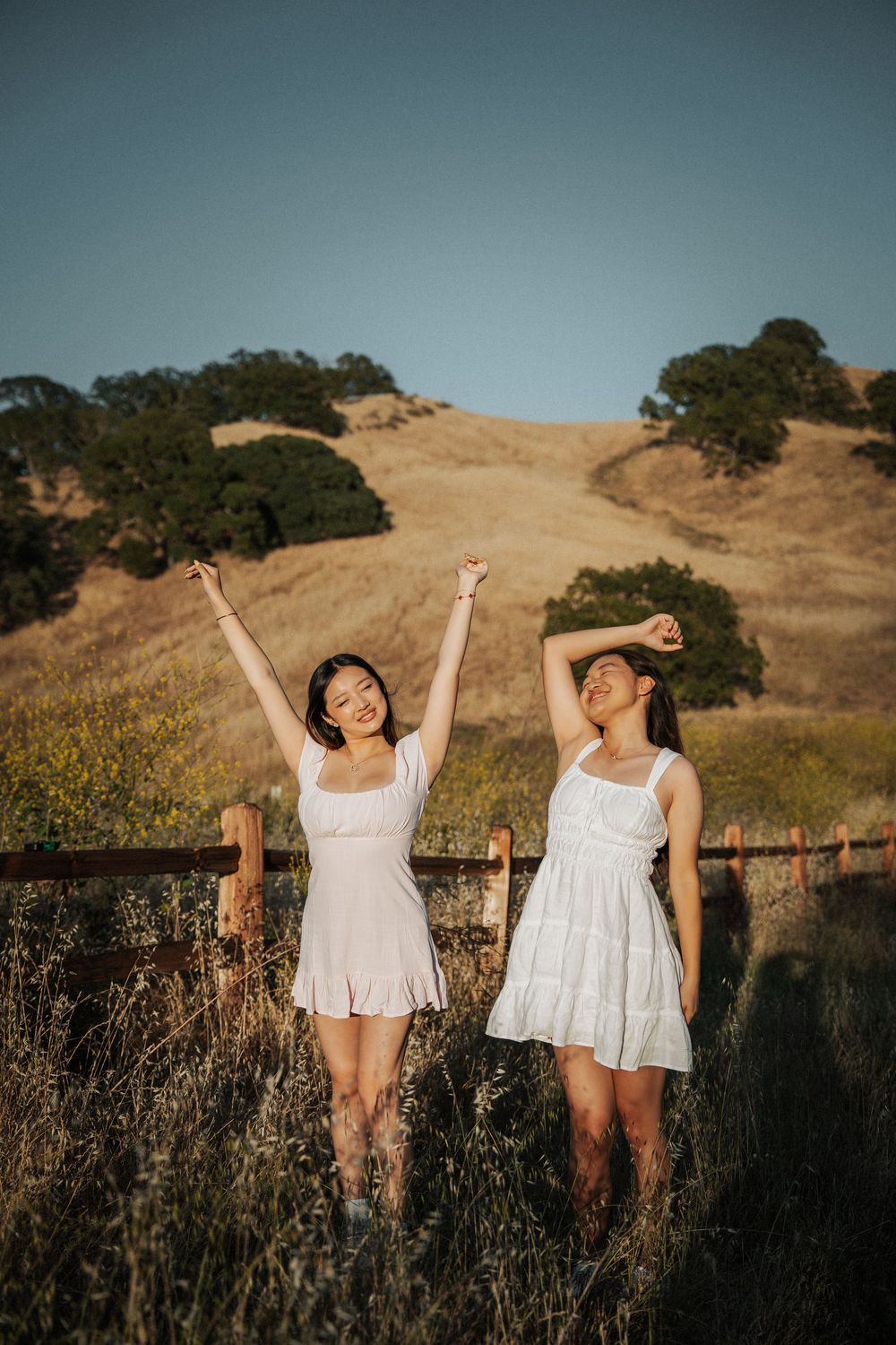 Two young women in white dresses celebrating with arms raised in a golden hillside meadow.