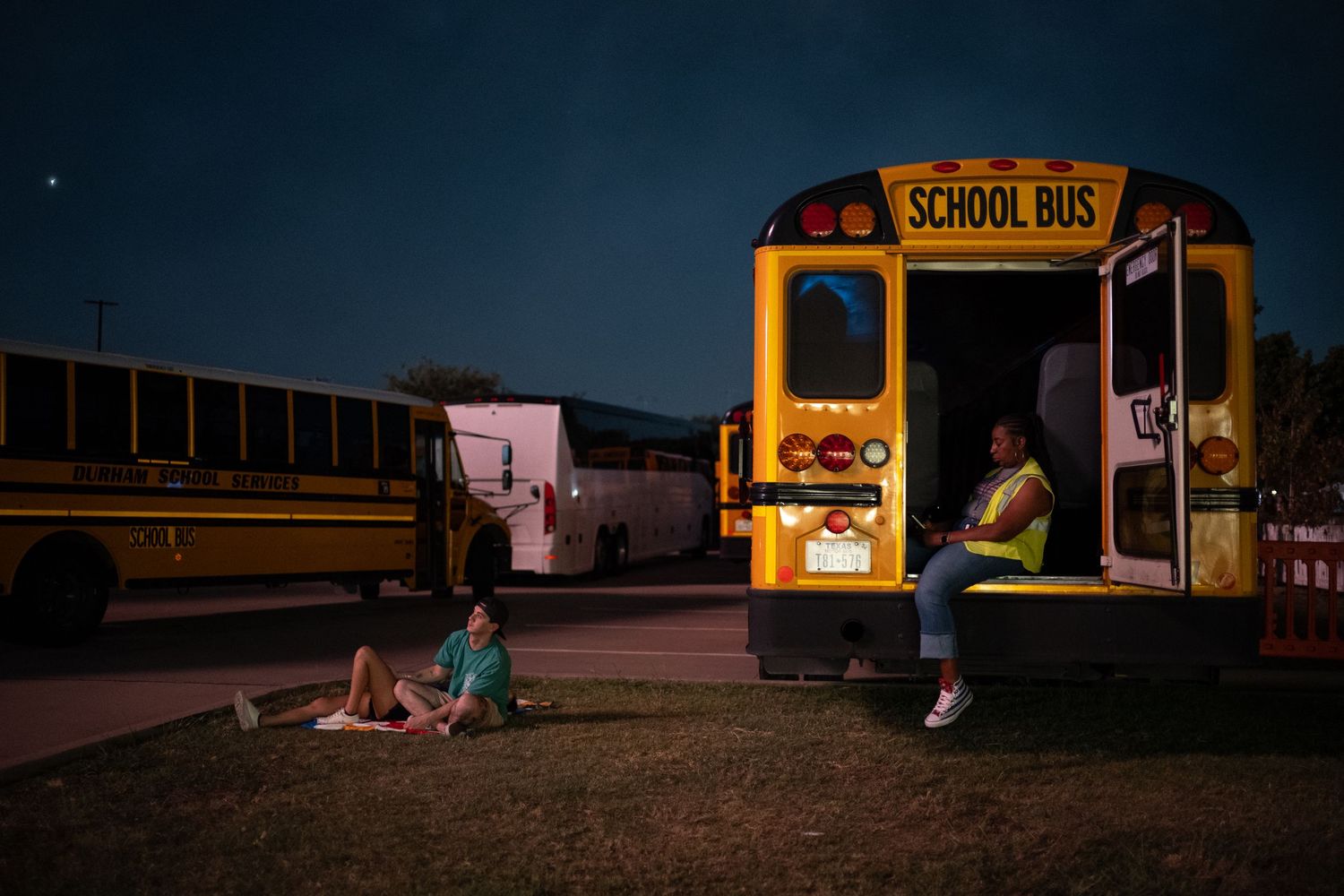 Two people relax near a yellow school bus at dusk, one sitting on the ground, another on the open door steps.