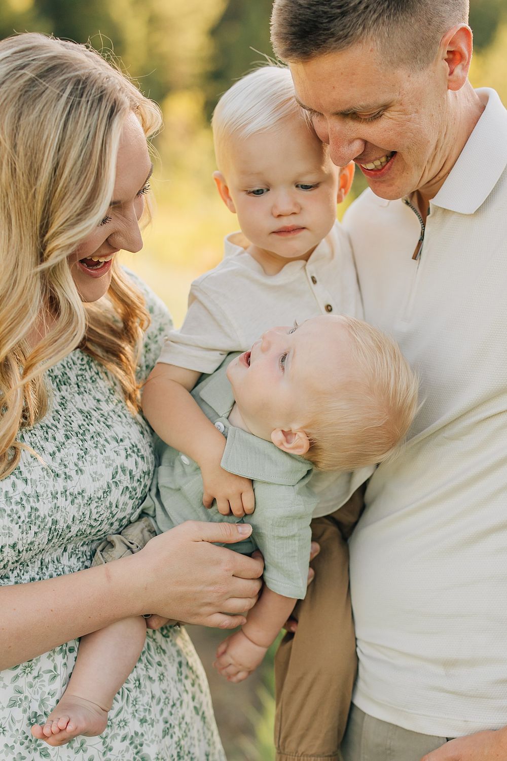 A happy family with two young blonde toddlers share a warm embrace outdoors in golden sunlight.