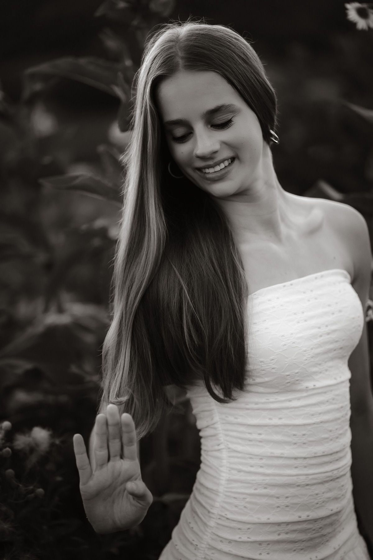 Black and white portrait of a smiling young woman with long hair in a white strapless dress, posing outdoors.
