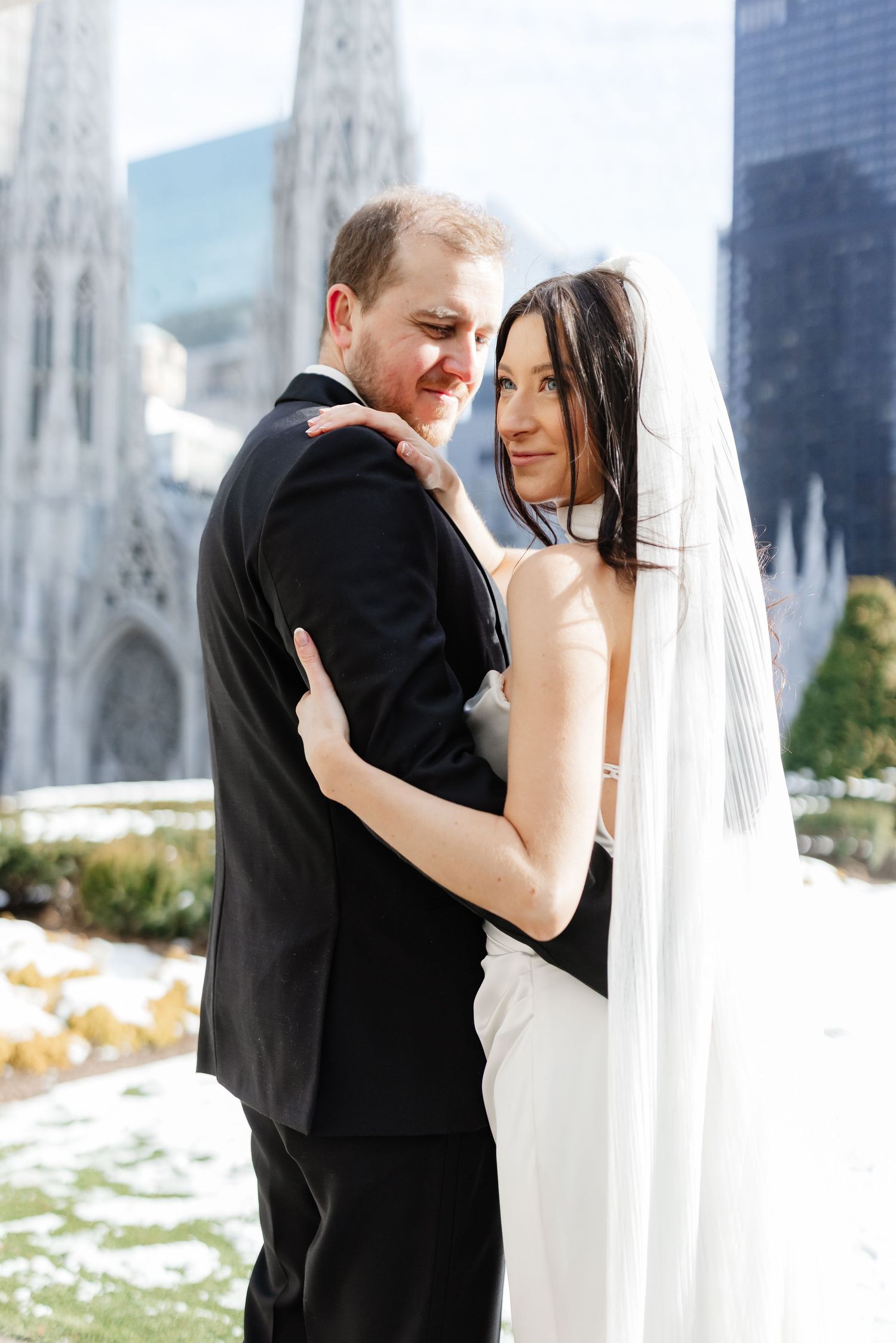 Bride and groom embrace outdoors in winter, with a Gothic cathedral and city skyline in the background.