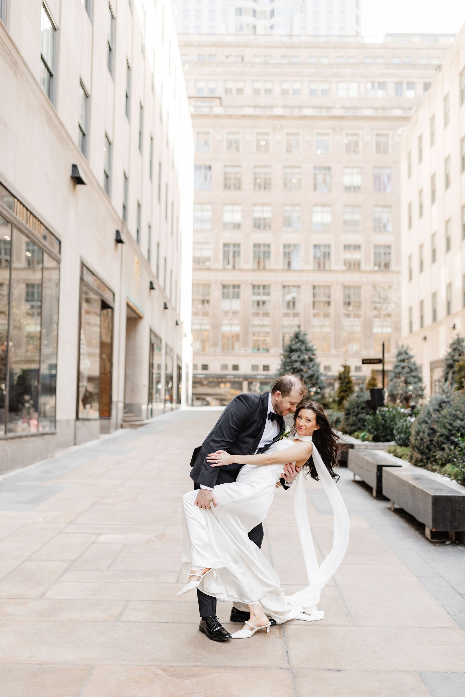 Couple shares a romantic dip pose in wedding attire on an urban sidewalk surrounded by elegant city buildings.