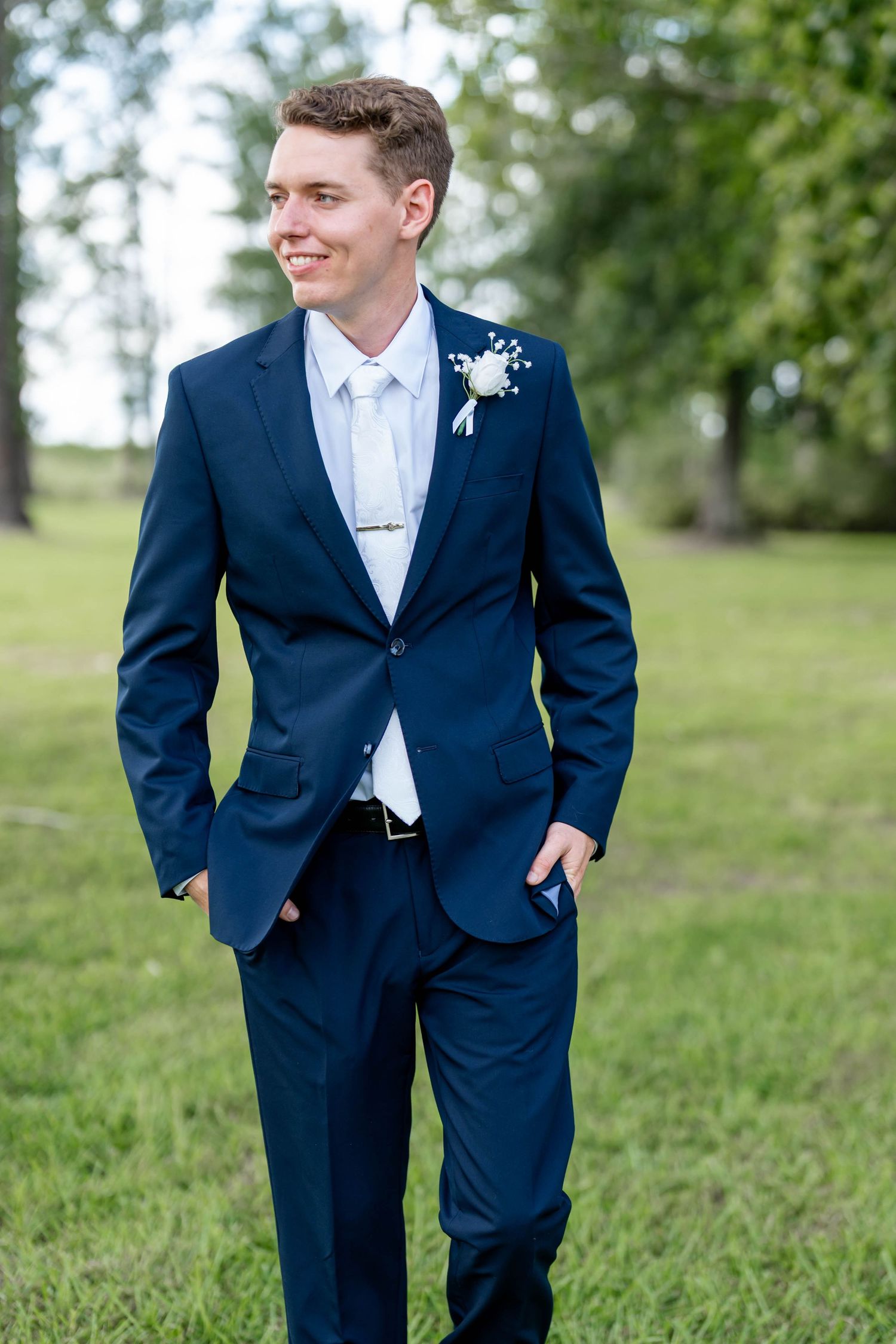 Young groom in navy blue suit with white boutonniere smiling outdoors on green grass at wedding.