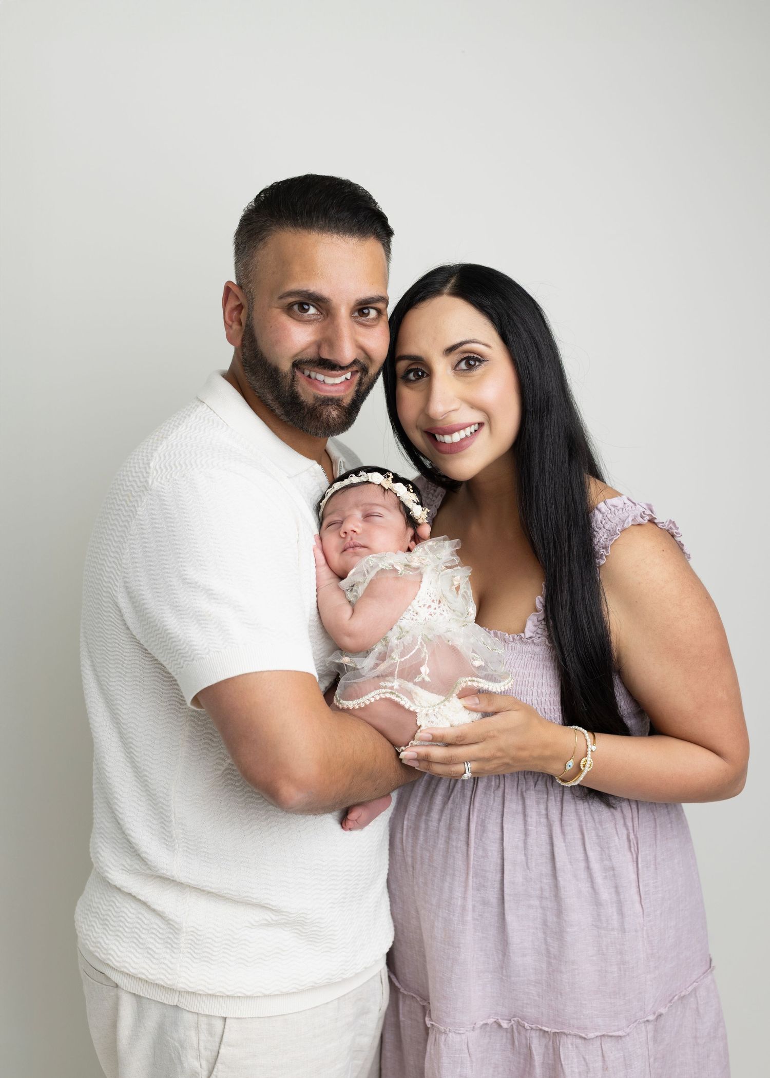 Smiling couple in a newborn photography session holding their sleeping baby dressed in white lace.