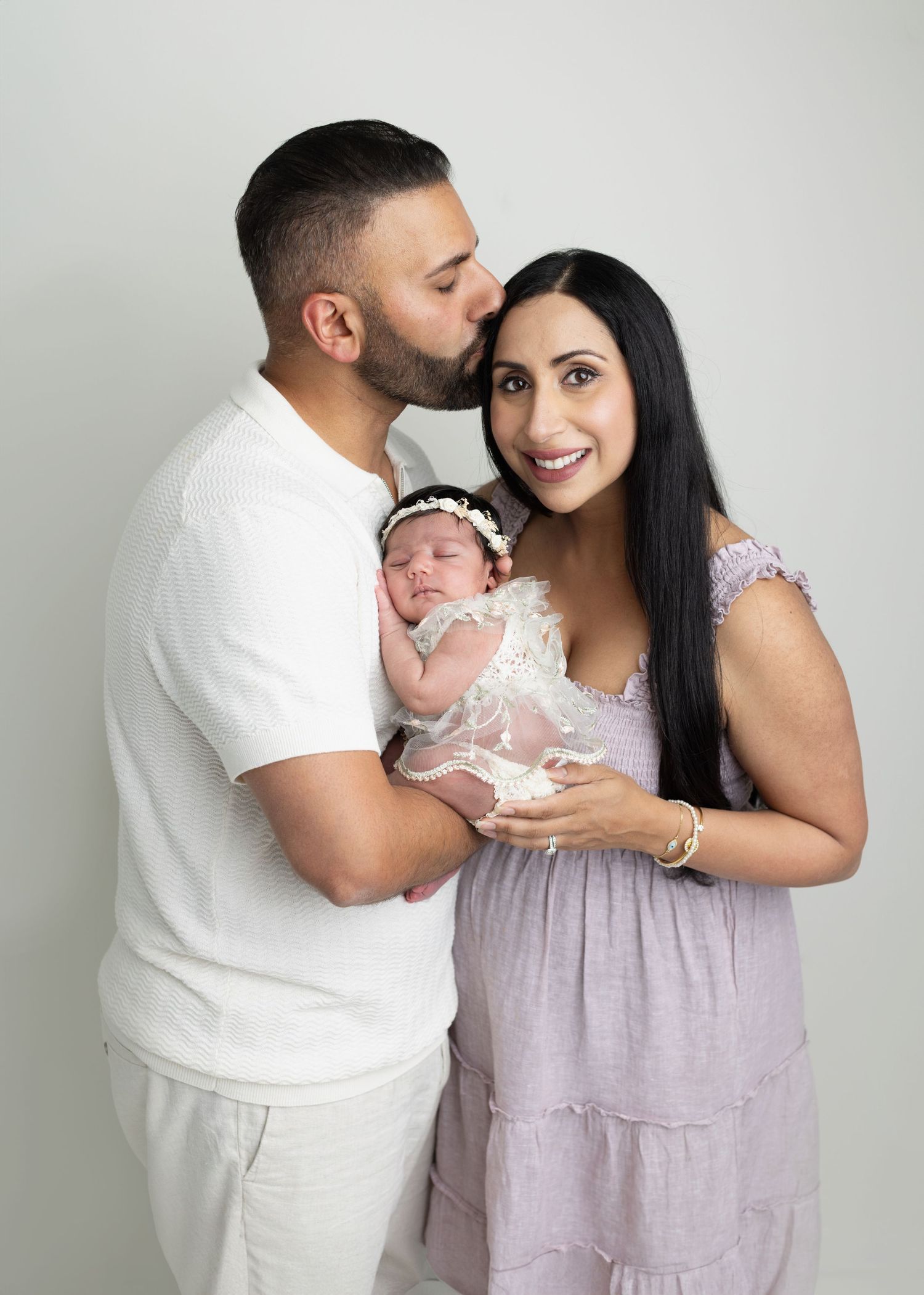 A couple smiles while holding their newborn baby dressed in lace, the man kissing the woman on the cheek.