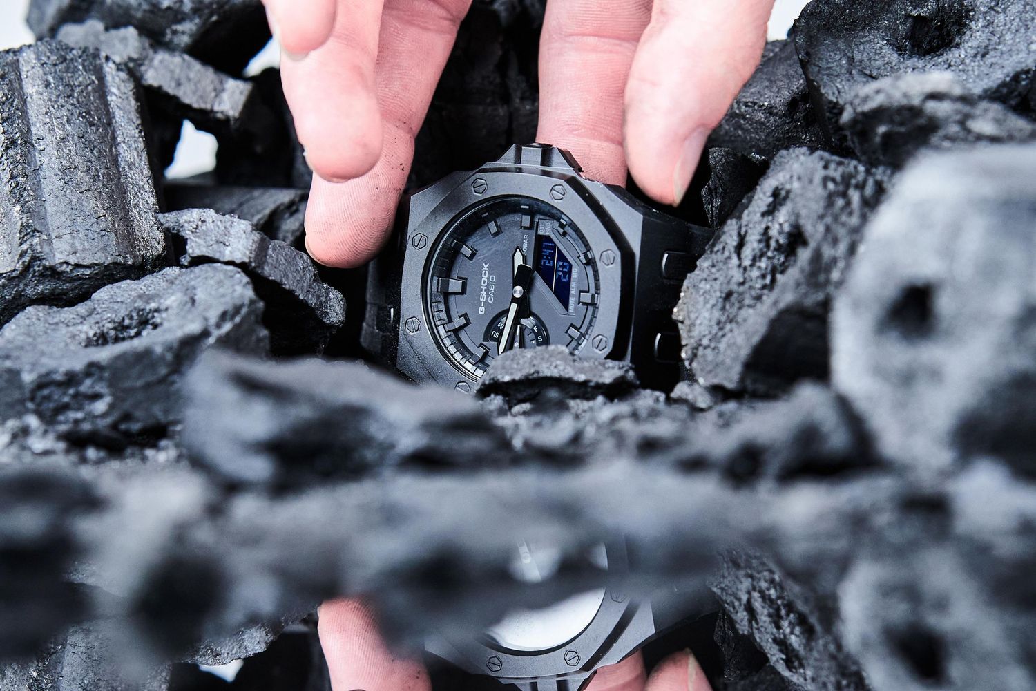 Hands holding a black G-Shock watch surrounded by dark charcoal pieces in a dramatic product photography setting.