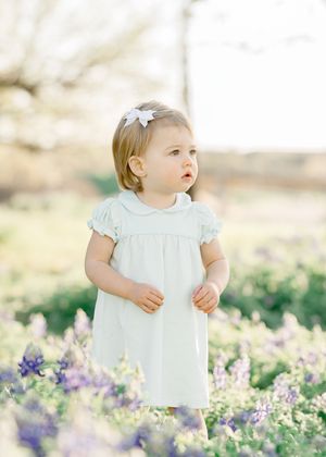 Toddler girl in mint dress and white bow stands among purple wildflowers in a sunlit field.