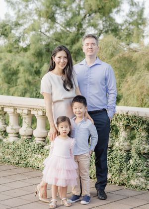 A family of four poses outdoors on a terrace, parents with two young children dressed in pastel outfits.