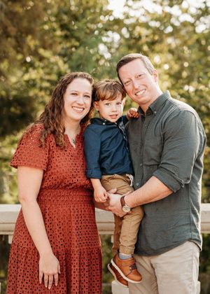 Happy family portrait of parents holding toddler son outdoors with lush green trees in background.
