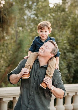 A smiling man carries a happy red-haired boy on his shoulders outdoors near a stone railing with lush greenery.