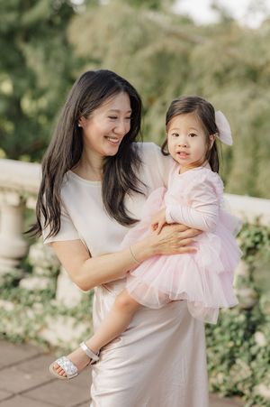 A smiling Asian woman in a white dress holds a toddler girl wearing a pink tutu dress with a bow outdoors.