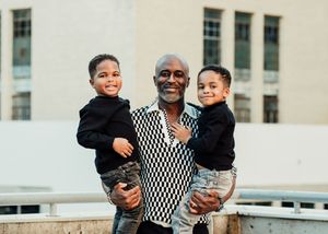 A man smiling and holding two young boys in his arms outdoors near a building.