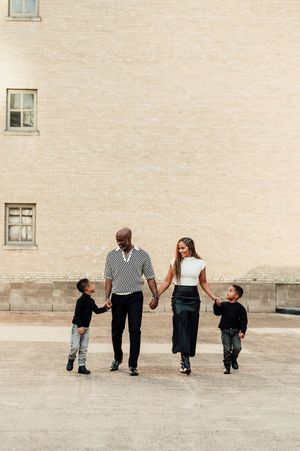 A joyful family of four walks hand-in-hand outdoors, with two young boys and their stylish parents near a brick building.