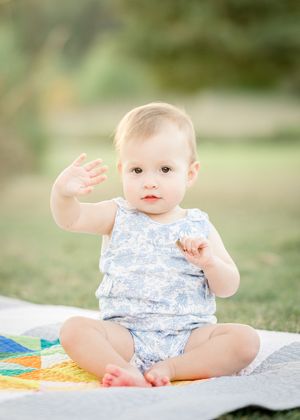 A cute baby in a blue floral romper sits on a colorful quilt outdoors, waving at the camera.