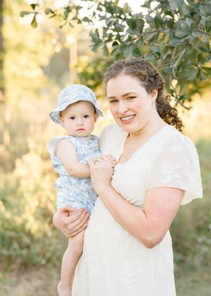 A smiling woman in a white dress holds a baby in a blue floral outfit and hat outdoors in natural light.