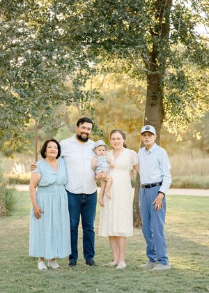 Multi-generational family of five poses outdoors under a large tree in soft natural light during a portrait session.