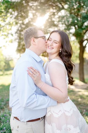 A couple shares a romantic moment outdoors, man kissing woman's cheek at golden hour in a sunlit park.