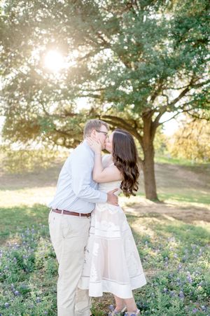 Couple sharing a romantic kiss outdoors under a large oak tree with golden sunlight filtering through the leaves.