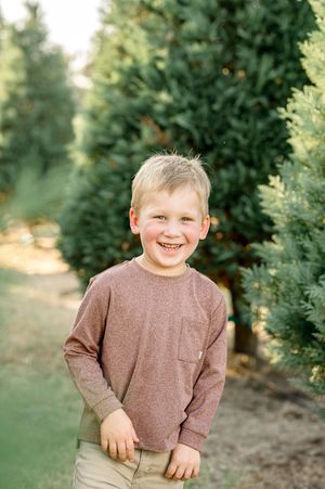 Smiling young boy with blonde hair wearing a mauve long-sleeve shirt standing among green trees at a Christmas tree farm.