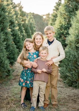 Family of four posing together at a Christmas tree farm, mother with three children smiling outdoors.