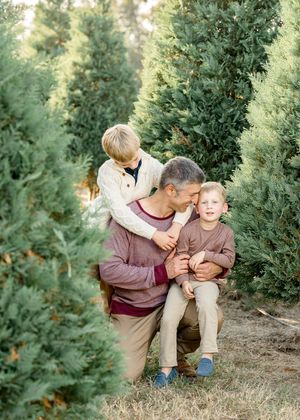 A father kneels with two young boys among evergreen trees at a Christmas tree farm.