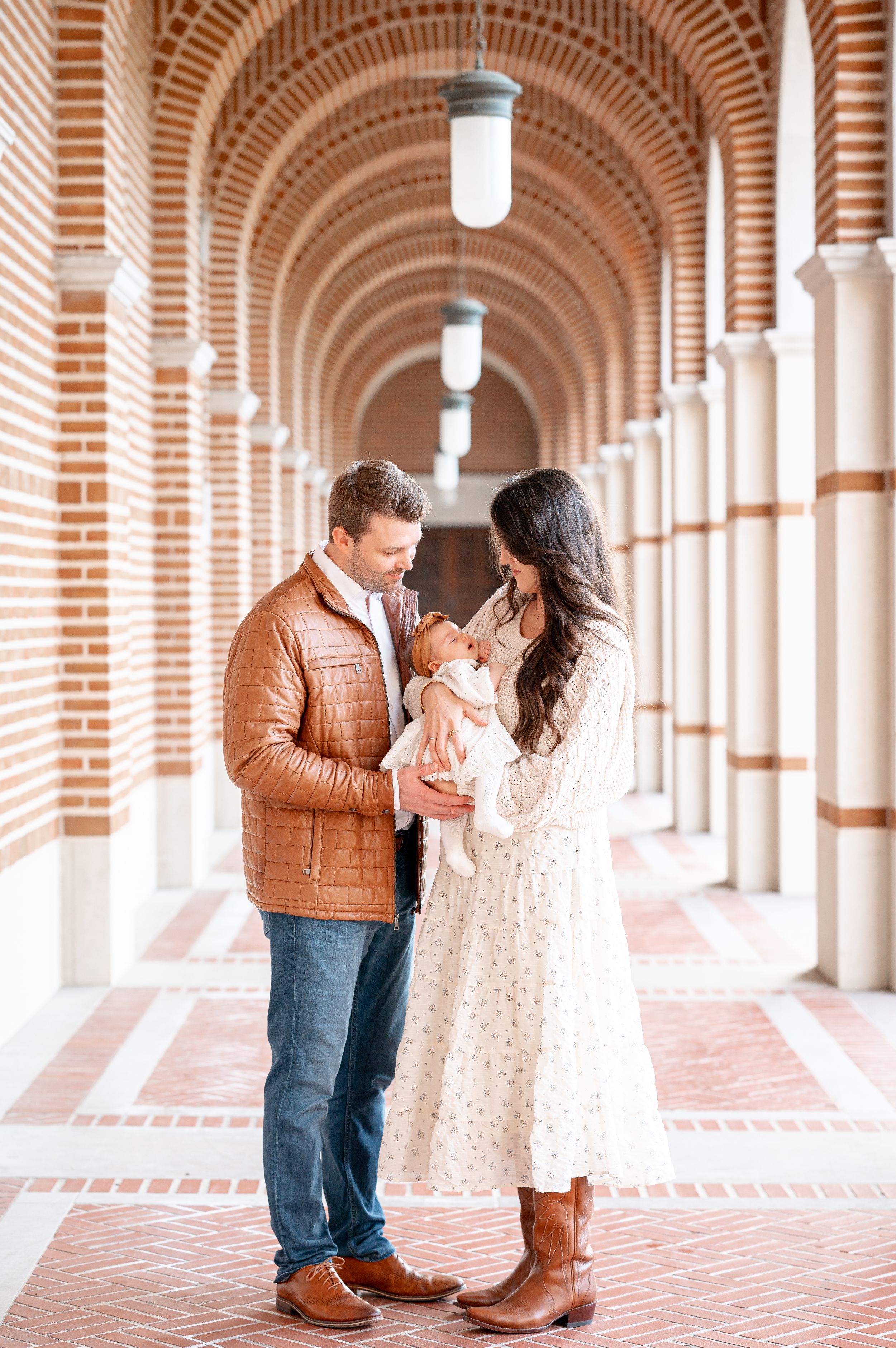 A couple admires their newborn baby in a elegant brick archway corridor, man in brown leather jacket and woman in floral dress.