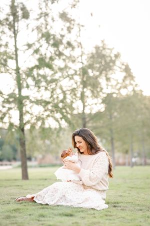 A mother sits on green grass cradling her baby outdoors, wearing a cream dress, surrounded by trees in soft golden light.