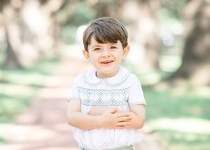 Smiling toddler boy in white smocked outfit posing outdoors on a tree-lined path for a portrait photo.