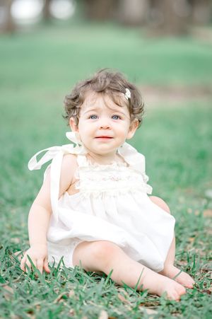 Adorable baby girl in white dress sitting on green grass outdoors, smiling with curly hair and a small bow.