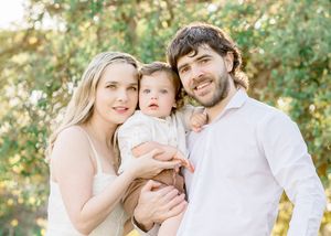 A smiling family of three poses outdoors, with parents holding their baby boy among lush green trees.