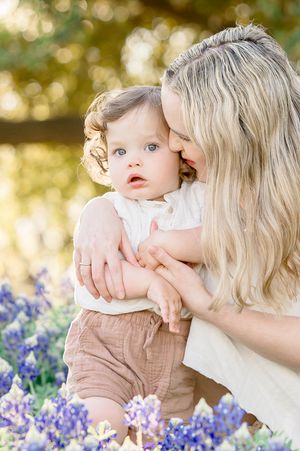 A mother kisses her baby among bluebonnets in a sunlit field, both wearing soft neutral tones.