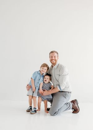A smiling father kneels beside two young children in a bright white studio family portrait.