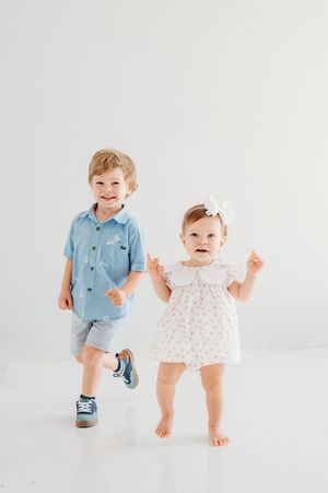 Two young children, a toddler boy in a blue shirt and shorts and a baby girl in a white floral dress, posing together.