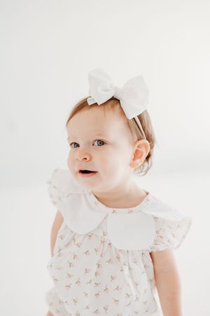 Adorable baby girl in a floral white romper with peter pan collar and white bow headband.