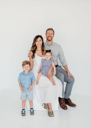 Happy family of four posing together in a bright white studio, parents with toddler son and baby daughter.