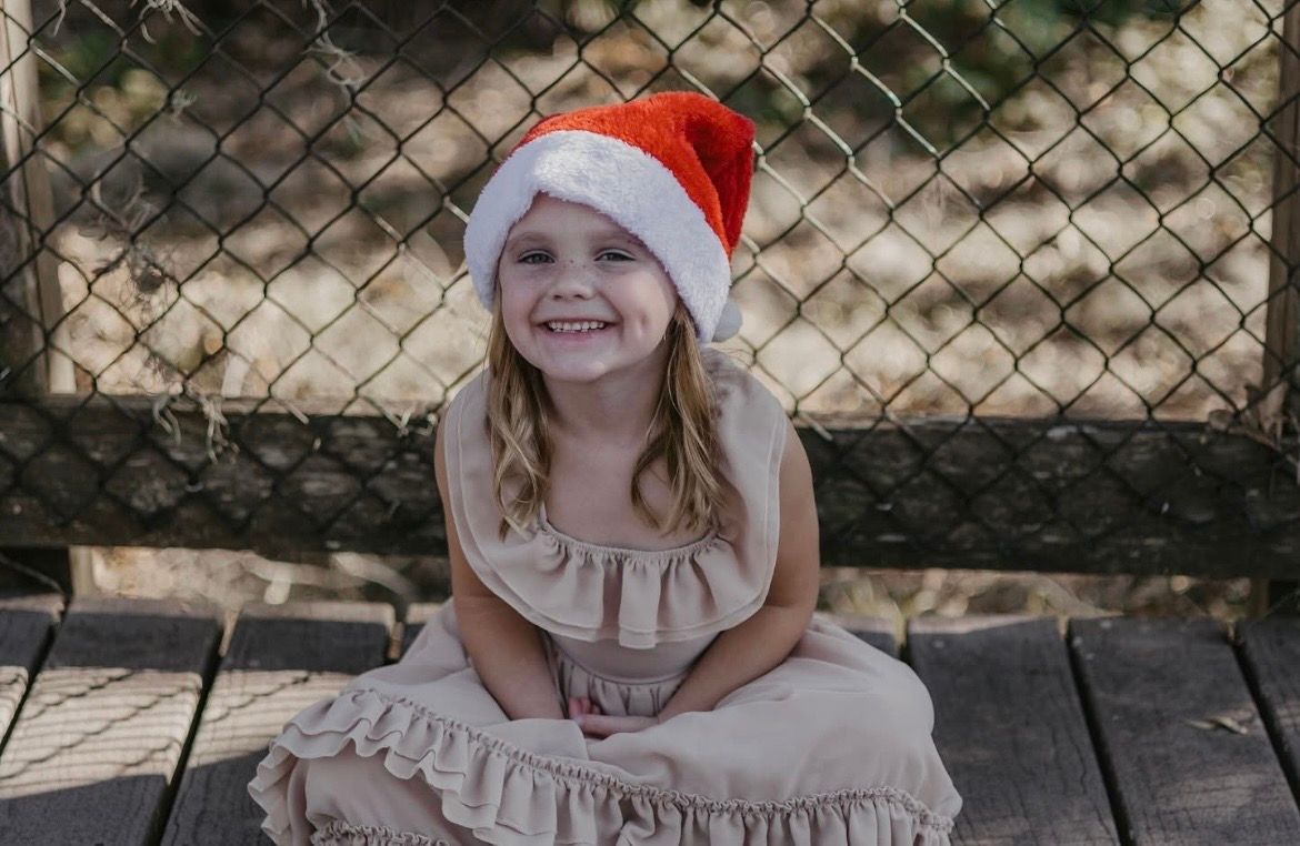 A smiling young girl wearing a Santa hat and beige dress sits on a wooden bench in front of a chain-link fence.